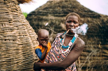 TOPOSA TRIBE, Güney Sudan - 12 Mart 2020: Toposa Kabilesinden bir kadın Güney Sudan, Afrika 'da geleneksel kulübelerin yanında dururken pipo içiyor ve bebek taşıyor