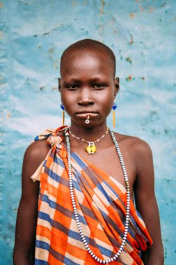 TOPOSA TRIBE, Güney Sudan - 12 Mart 2020: Kareli renkli elbiseli ve geleneksel aksesuarlı kız Güney Sudan 'daki Toposa Kabile Köyü' nde mavi duvara bakan kameraya bakıyor