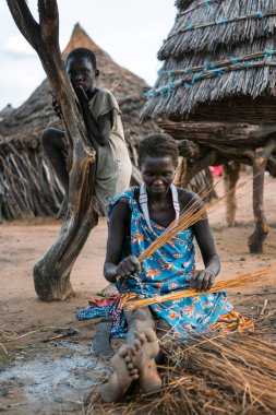 TOPOSA TRIBE, Güney Sudan - 12 Mart 2020: Güney Sudan, Afrika 'da Toposa Kabilesi köyünde baraka ve çocuğun yanındaki kirli zeminde oturan ve samandan ürün dokuan kadın