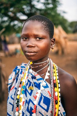 TOPOSA TRIBE, Güney Sudan - 12 Mart 2020: Süslü giysili ve renkli aksesuarlı genç kız Güney Sudan 'daki Toposa Kabile Köyü' nün bulanık arka planında kameraya bakıyor