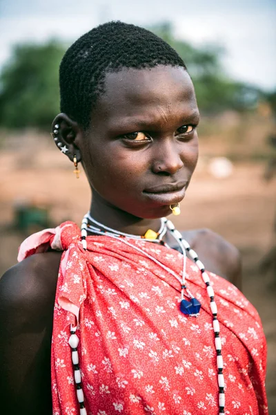 TOPOSA TRIBE, SOUTH SUDAN - 12 maart 2020: Tiener met kort haar in helder kledingstuk en accessoires en kijkend naar camera op wazige achtergrond van Toposa Tribe dorp in Zuid-Soedan, Afrika — Stockfoto