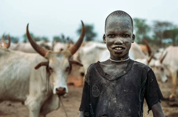 MUNDARI TRIBE, SOUTH SUDAN - MARCH 11, 2020: Boy shepherd in rags smiling at camera with interest while standing against herd of cows with big and sharp horns on pasture in South Sudan in daylight — Stock Photo, Image