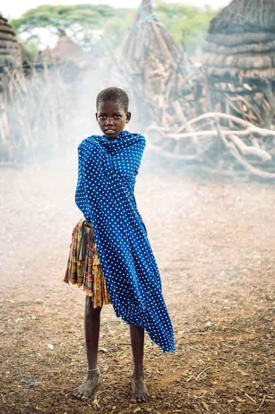 TOPOSA TRIBE, SOUTH SUDAN - MARCH 12, 2020: Kid in skirt wrapping in dotted blue fabric against smoke and huts in Toposa Tribe village in South Sudan, Africa — Stock Photo, Image