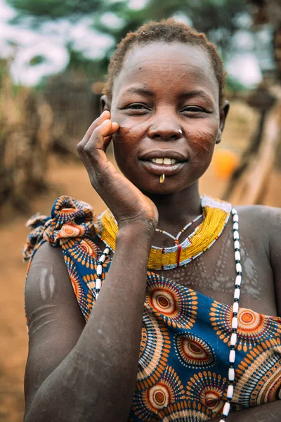 TOPOSA TRIBE, SUD SUDAN - 12 MARZO 2020: Giovane donna con le tradizionali cicatrici della Tribù Toposa che tocca il viso e guarda la fotocamera sullo sfondo sfocato del villaggio in Sud Sudan, Africa — Foto Stock