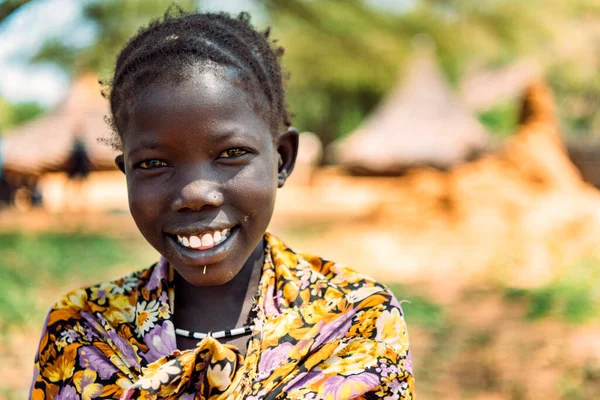 BOYA TRIBE, SÜDSUDAN - 10. MÄRZ 2020: Mädchen in traditionellem, farbenfrohem Outfit und Accessoire lächelt vor verschwommener Siedlung im Südsudan in Afrika in die Kamera lizenzfreie Stockfotos