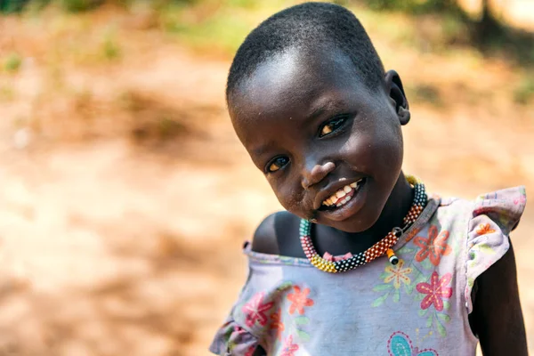 BOYA TRIBE, SOUTH SUDAN - MARCH 10, 2020: Small girl in colorful dress and traditional necklace smiling at camera against blurred rural environment in South Sudan in sunny day Stock Image