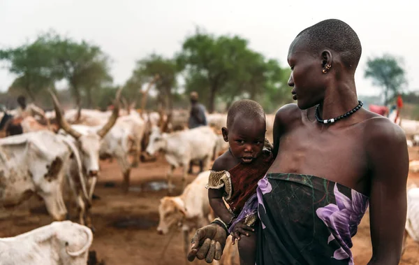 MUNDARI TRIBE, SUDÃO DO SUL - MARÇO 11, 2020: Mulher com roupas e acessórios tradicionais carregando bebê curioso a mãos enquanto está de pé contra o ambiente rural turvo e rebanho de vacas no Sul Imagem De Stock