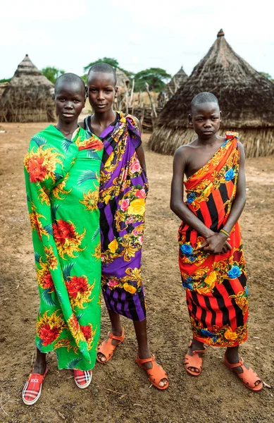 TOPOSA TRIBE, SOUTH SUDAN - 12 maart 2020: Tieners gewikkeld in kleurrijke, heldere stof kijken naar camera tegen strohutten in het dorp Toposa Tribe in Zuid-Soedan, Afrika Rechtenvrije Stockafbeeldingen