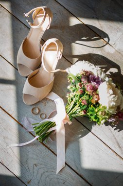 Düğün için. Dekorasyon. Gelinin ayakkabıları, güzel bir düğün buketi, yüzükler, yaka çiçekleri ahşap bir arka planda güzelce serilmiş. Üst Manzara. Gölgeler. Güneş ışığı.