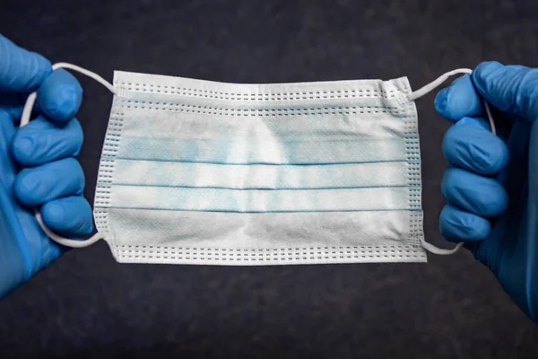 Tıbbi ya da cerrahi açık mavi maske yakın plan elinde koyu renkli halı tekstil arka planında mavi eldivenlerle. Kişisel koruma için tıbbi malzeme veya aksesuar kavramı.