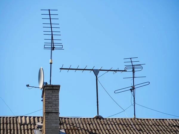 Satellite and old television antennas are installed on the sloping slate roof of the house. Antennas are attached with cables. Rooftop chimney. Against the backdrop of a clean blue sky.