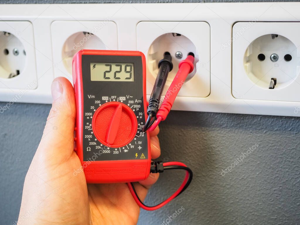 Measuring electricity in a socket using a multimeter in the hand. Red pocket digital multimeter with LCD display. Test leads in the socket. High-quality Close-up photography.