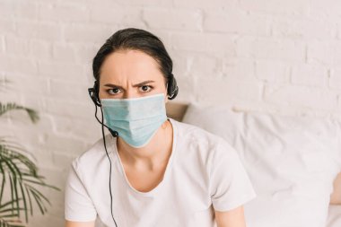 Evde çalışırken kameraya bakan kulaklık ve tıbbi maske takan ciddi bir uzaktan kumandacı.