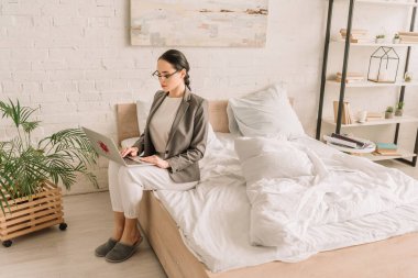 young businesswoman in blazer over pajamas using laptop while sitting on bed clipart