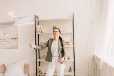 Pijama giyip yatak odasındaki kameraya bakarken dokümanları kusan spor ceketli mutlu bir iş kadını.