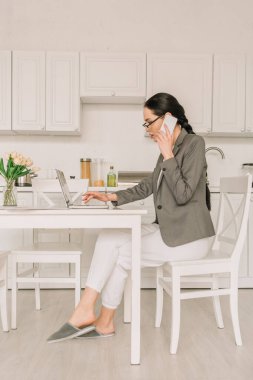 side view of businesswoman in blazer over pajamas working in kitchen, talking on smartphone and using laptop clipart