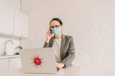 Tıbbi maskeli ve gözlüklü ciddi bir iş kadını diz üstü bilgisayarın yanında Coronavirus bakteri çıkartmalı akıllı telefonla konuşuyor.