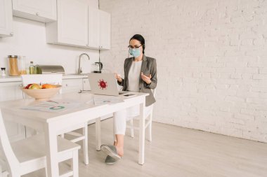 Medikal maskeli ve spor ceketli bir iş kadını, dizüstü bilgisayarın yanında çalışırken pijama giyerken el hareketi yapıyor.