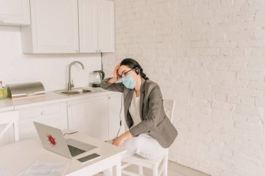 Pijama, tıbbi maske ve kulaklık takmış spor ceketli yorgun bir iş kadını evde çalışırken dizüstü bilgisayarın yanında oturuyor.
