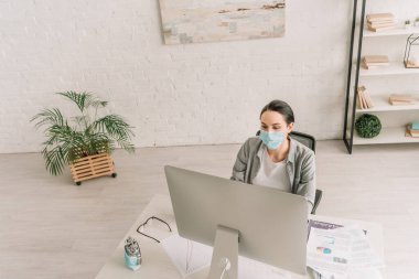 Medikal maskeli serbest çalışanın evde bilgisayar monitörünün yanında çalışmasının yüksek açılı görüntüsü