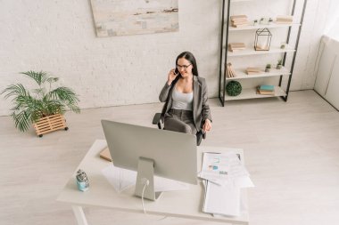 Yüksek açılı iş kadını masadaki akıllı telefonla konuşurken bilgisayar monitörü ve tıbbi maskeye sarılı çalar saatle görülüyor.