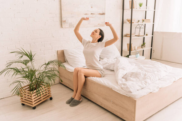Улыбающаяся женщина в пижаме, растянутая с закрытыми глазами, сидя на кровати рядом с растением в горшке
