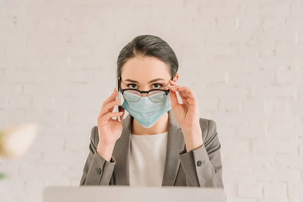 Tıp maskeli ve gözlüklü ciddi bir iş kadını evde akıllı telefondan konuşuyor.