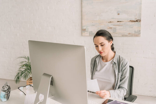 young, attentive freelancer looking at document while sitting near computer monitor