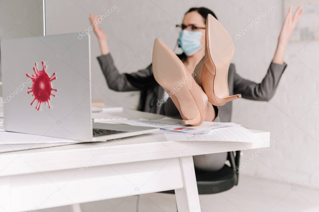 selective focus of businesswoman in medical mask showing shrug gesture while working at home with legs on table