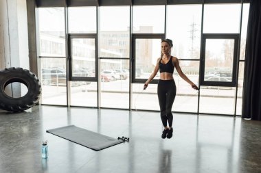 Sporcu kadın halatla atlıyor fitness minderinin yanında halat atlıyor, halterler ve spor şişesi spor salonunda.