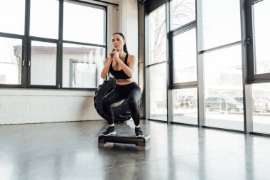 Sporcu kadın spor merkezinde adım platformunda çömeliyor.
