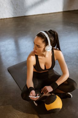Yüksek açılı, kulaklıklı çapraz bacaklı sporcu kadın spor salonundaki spor minderinin yanında akıllı telefon tutuyor.