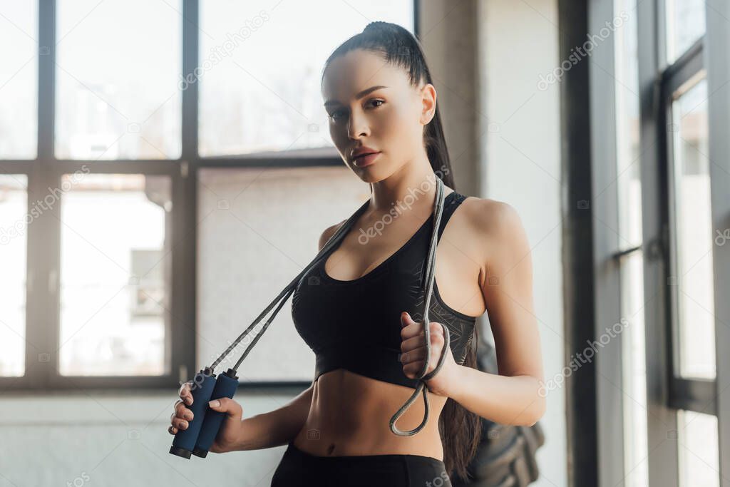Beautiful sportswoman holding skipping rope and looking at camera in gym