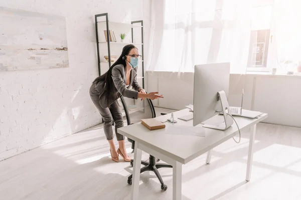 Joven mujer de negocios en desgaste formal y máscara médica que apunta con el dedo al monitor de la computadora en casa - foto de stock