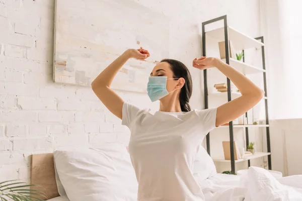 Mujer joven en pijama y máscara médica estirando las manos en el dormitorio - foto de stock