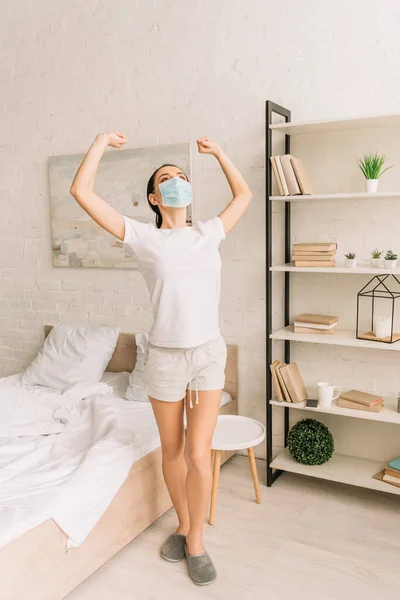 Jeune femme en pyjama et masque médical étirant les mains dans la chambre — Photo de stock