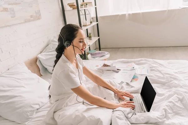 Side view of remote operator in headset typing on laptop while working in bed — Stock Photo
