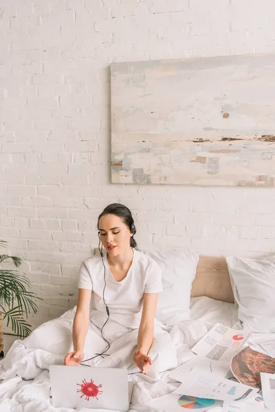 Junge Fernbedienung in Headset arbeitet im Bett in der Nähe von Laptop mit Coronavirus-Bakterien Aufkleber — Stockfoto