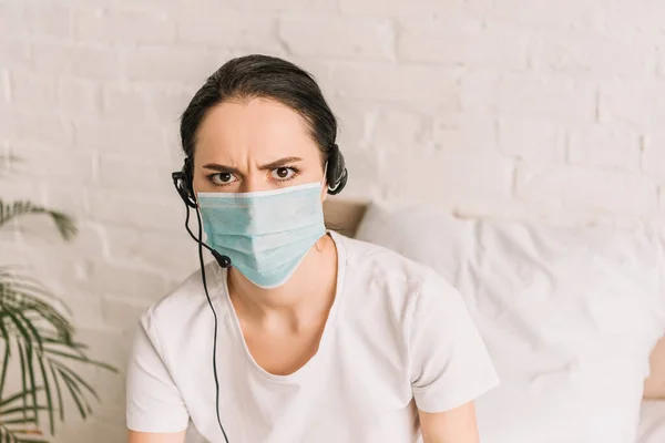 Serious remote operator in headset and medical mask looking at camera while working at home — Stock Photo