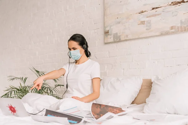 Operador remoto en máscara médica y auriculares apuntando con el dedo a la pantalla del ordenador portátil mientras trabaja en la cama - foto de stock