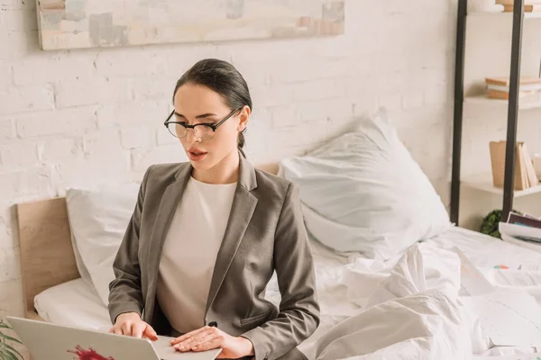 Beautiful businesswoman in blazer over pajamas using laptop in bedroom — Stock Photo