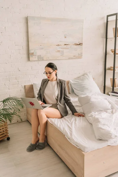 Attractive businesswoman in blazer over pajamas using laptop while sitting on bed — Stock Photo