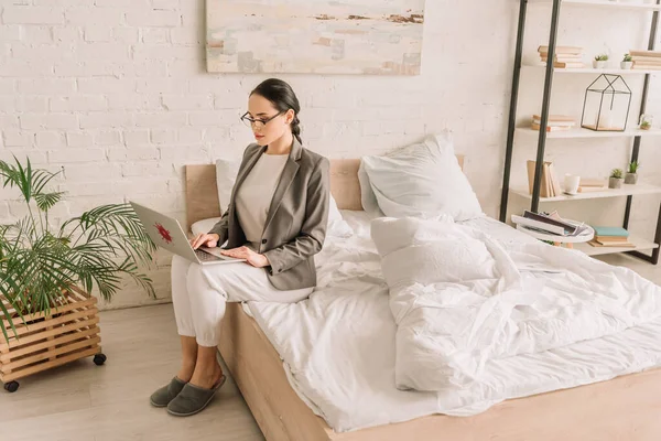 Jeune femme d'affaires en blazer sur pyjama à l'aide d'un ordinateur portable tout en étant assis sur le lit — Photo de stock