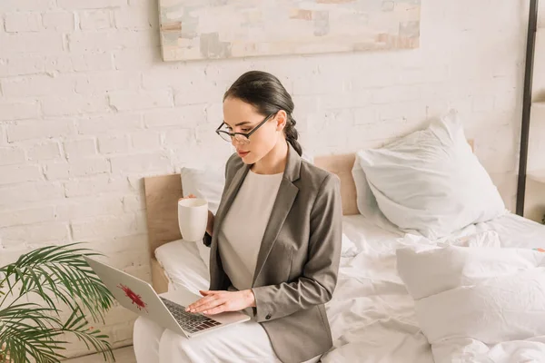 Attentive businesswoman in blazer over pajamas using laptop and holding cup of coffee in bedroom — Stock Photo