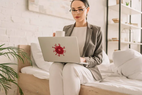 Attractive businesswoman in blazer over pajamas using laptop and holding cup of coffee while sitting on bed — Stock Photo