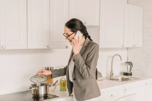 Smiling businesswoman in blazer over pajamas cooking and talking on smartphone in kitchen — Stock Photo