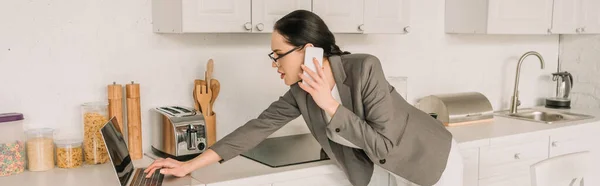 Cultivo panorámico de mujer de negocios en blazer sobre pijamas utilizando el ordenador portátil y hablando en el teléfono inteligente en la cocina - foto de stock