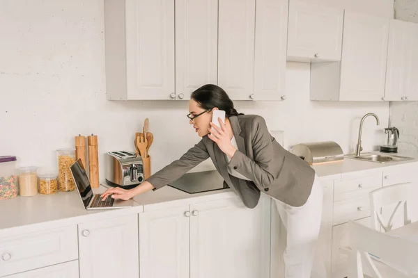 Mujer de negocios en blazer sobre pijamas hablando en el teléfono inteligente y el uso de ordenador portátil en la cocina - foto de stock