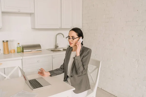 Young businesswoman in blazer over pajamas talking on smartphone while working in kitchen near laptop — Stock Photo