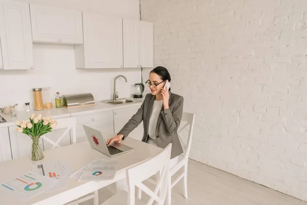 Улыбающаяся бизнесвумен в пижаме разговаривает на смартфоне и пользуется ноутбуком на кухне рядом с документами и свежими тюльпанами — стоковое фото